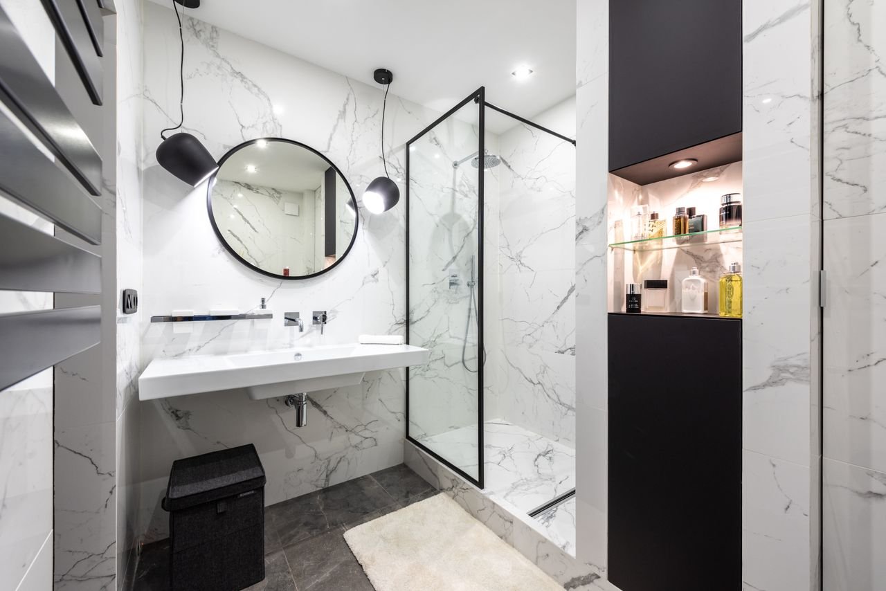 Shower room against washbasin and mirror between shiny lights in bathroom with assorted perfume in bottles on shelves